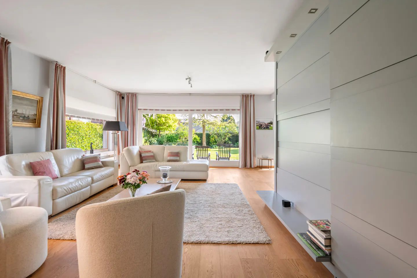 Bright living room with white sofas, a beige rug, and a large window overlooking a green garden. A modern gray wall unit is on the right.