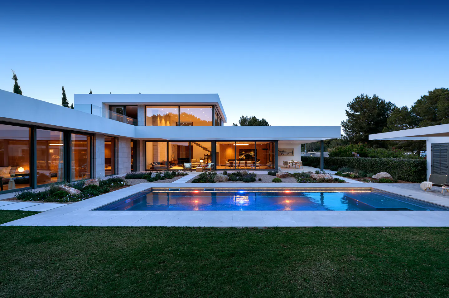 Modern white house with large windows and a blue-lit pool at dusk. Green lawn in the foreground.