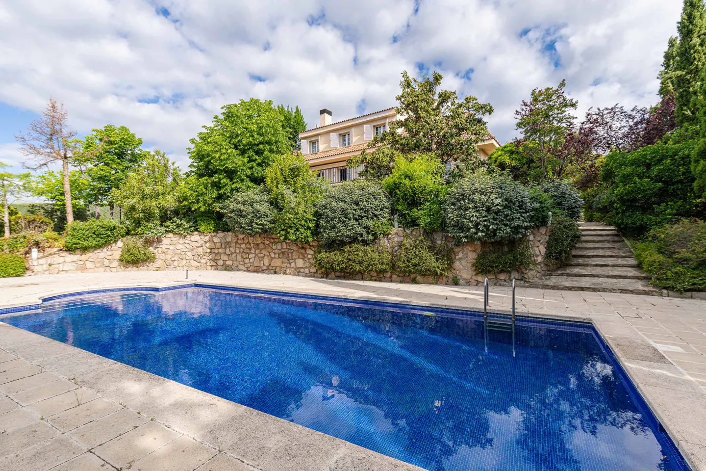 A blue tiled swimming pool in front of a beige house with green trees and a stone wall.