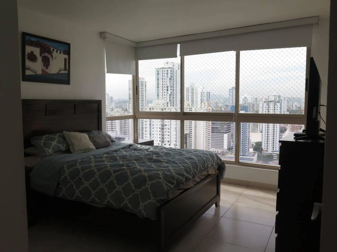 Bedroom with a queen bed, blue patterned comforter, and a large window overlooking a city skyline. A TV sits on a dresser.