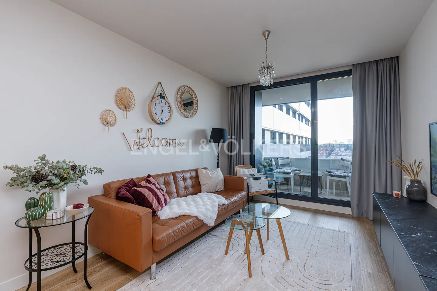 Modern living room with a tan leather sofa, glass coffee tables, and a wall decorated with a "Welcome" sign and clock.