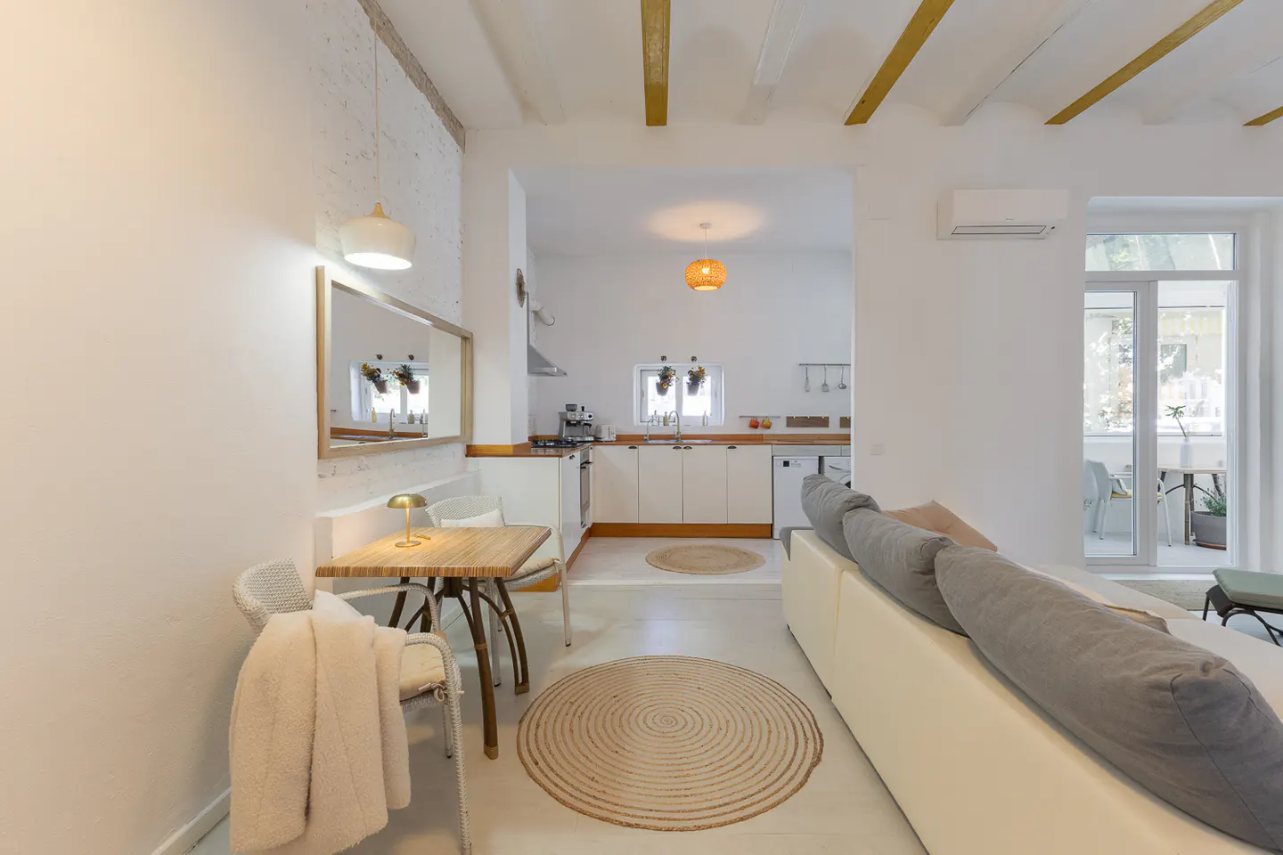 Bright, open-concept living space with white walls, wooden beams, and a jute rug. A small dining table sits near a mirror, and a kitchen is visible in the background.