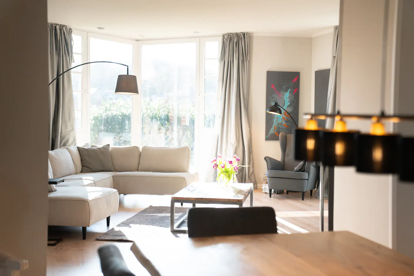 Bright living room with a beige sectional sofa, grey armchair, coffee table with flowers, and large windows with curtains.