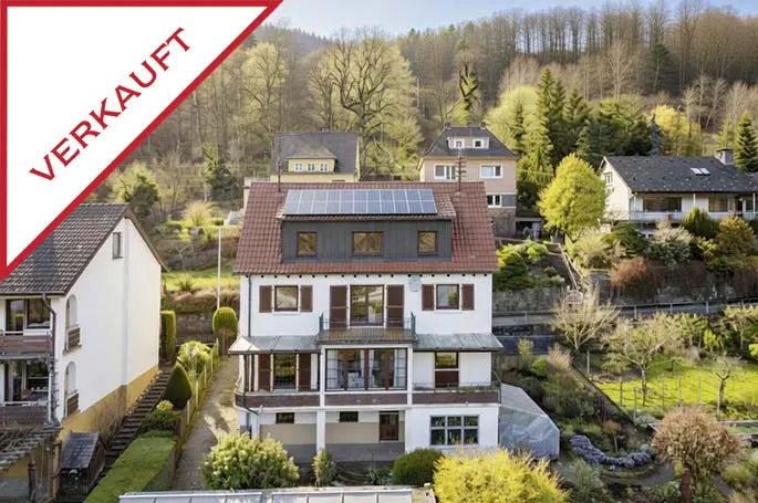 A three-story white house with a red roof and solar panels, surrounded by trees. A red "VERKAUFT" banner is in the upper left corner.