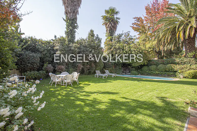 Lush green backyard with a pool, lounge chairs, and a white table with chairs. Engel & Volkers logo in the background.