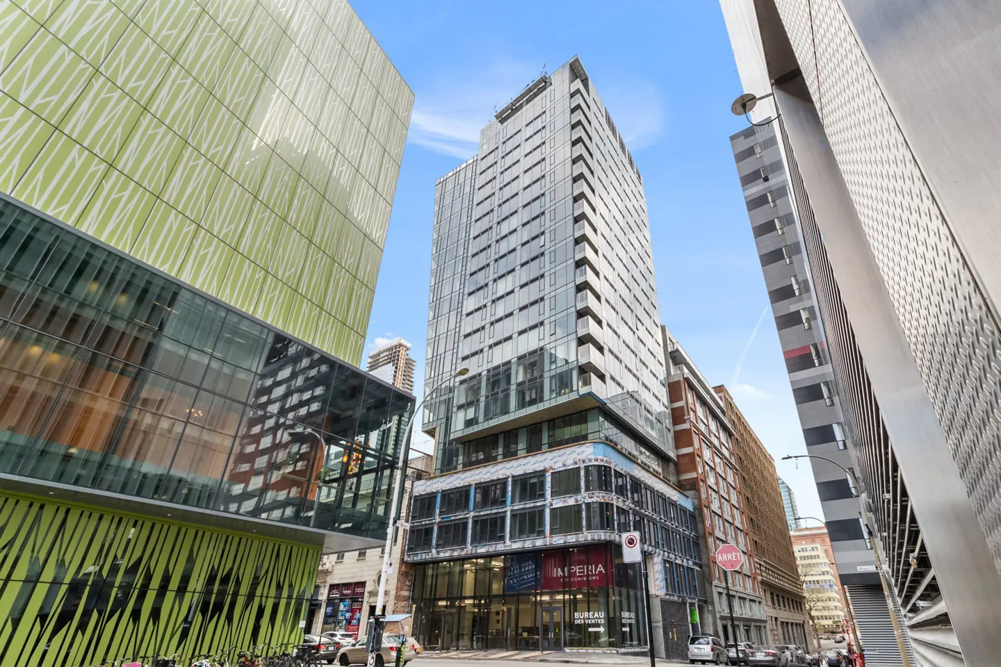 Street-level view of a tall, modern glass building in a city. Other buildings are visible, including one with green patterned panels.