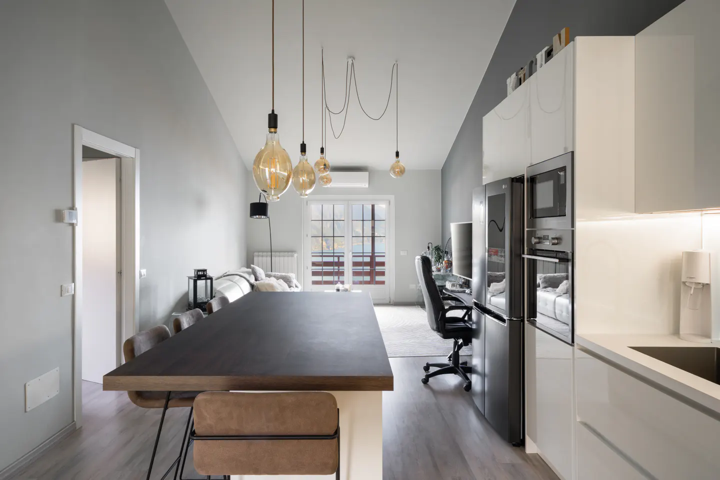 Open-concept living space with a kitchen island, dining area, and lounge. Modern lighting fixtures hang above a dark wood table with brown chairs. Balcony doors offer a view of the water.