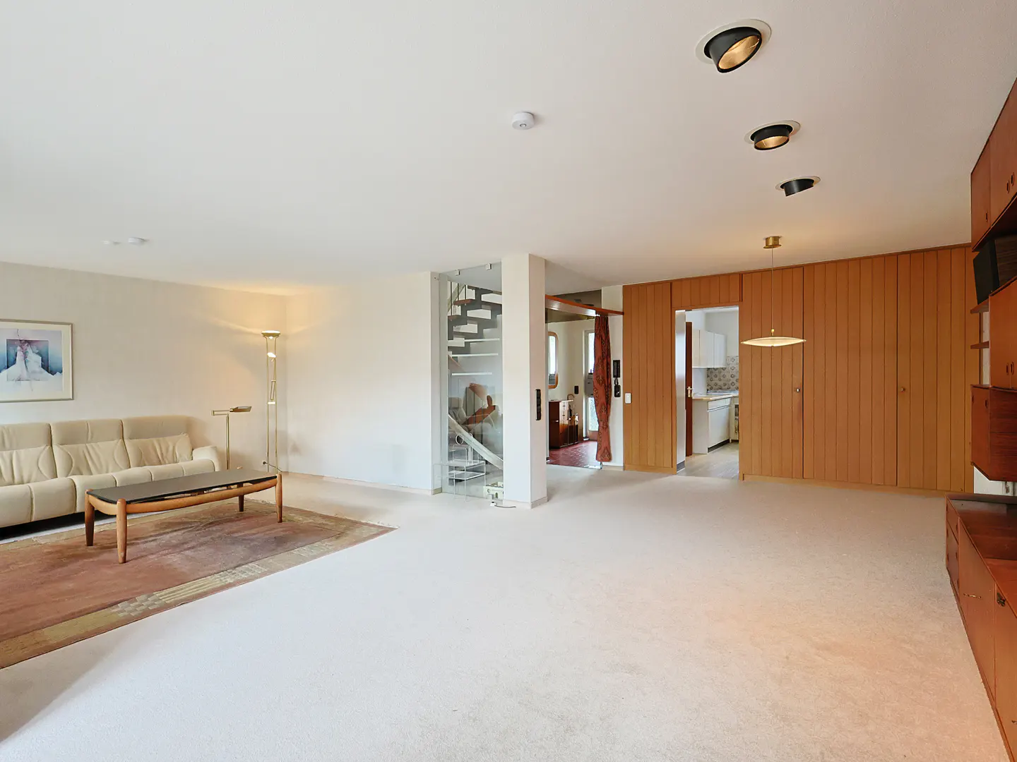 Bright, open living room with beige sofa, wood table, and spiral staircase. Wood paneled wall with recessed lighting and kitchen view.