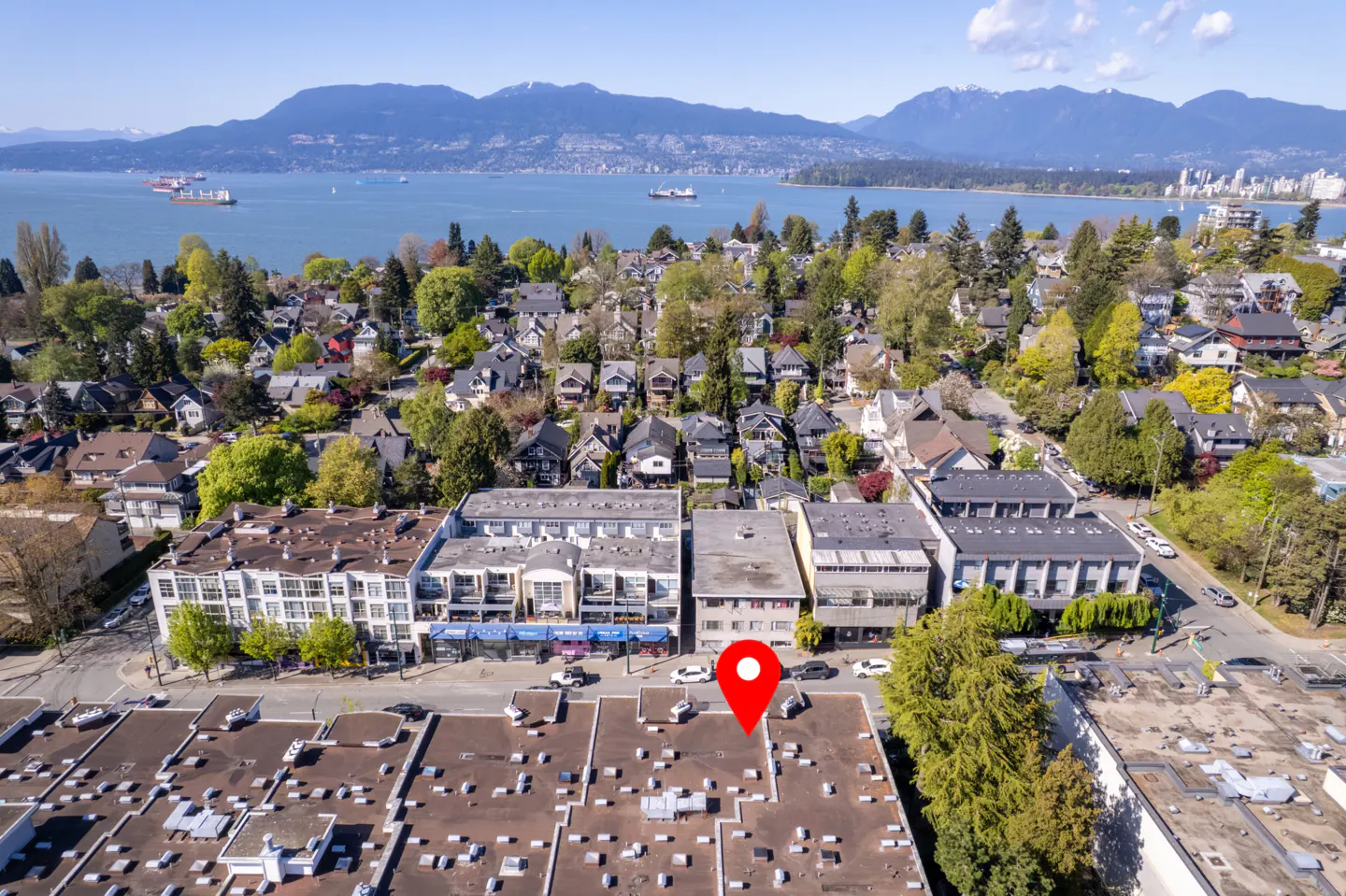 Aerial view of a city neighborhood with a red location pin on a building roof. Mountains and water are in the background.