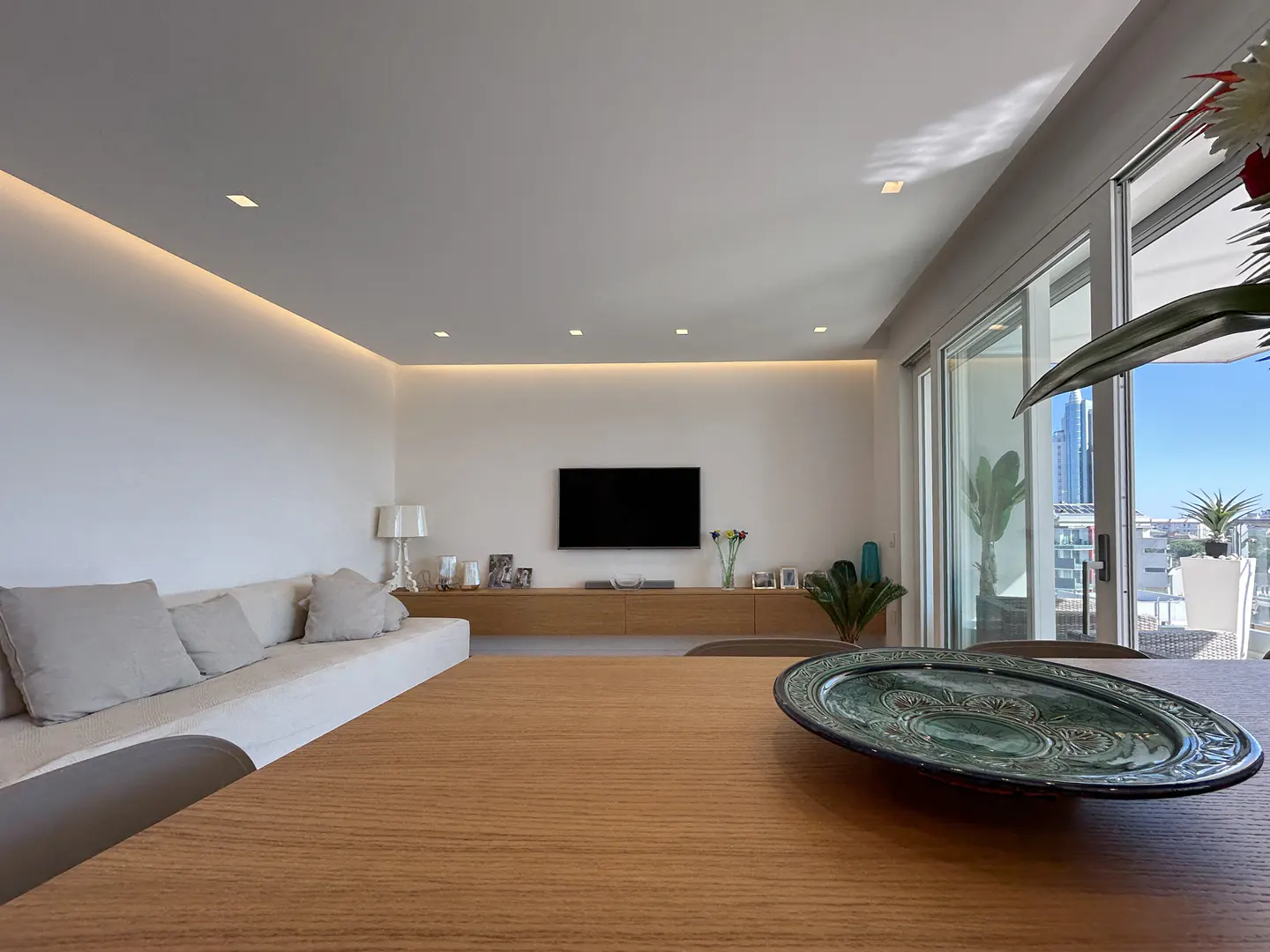 Bright, modern living room with a wood table, white sofa, and a large TV. Sliding glass doors lead to a balcony with city views.