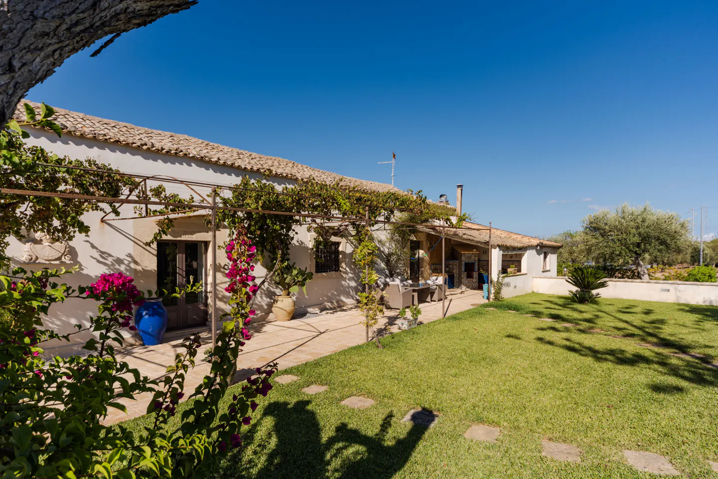 Exterior of a white, one-story house with a tile roof, green lawn, and blue sky. A pergola covered in vines runs along the front of the house.