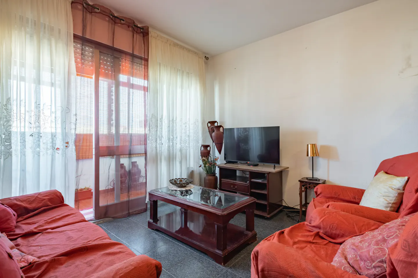 A living room with red sofas, a glass-topped coffee table, and a TV on a wooden stand. Curtains hang by a balcony door.