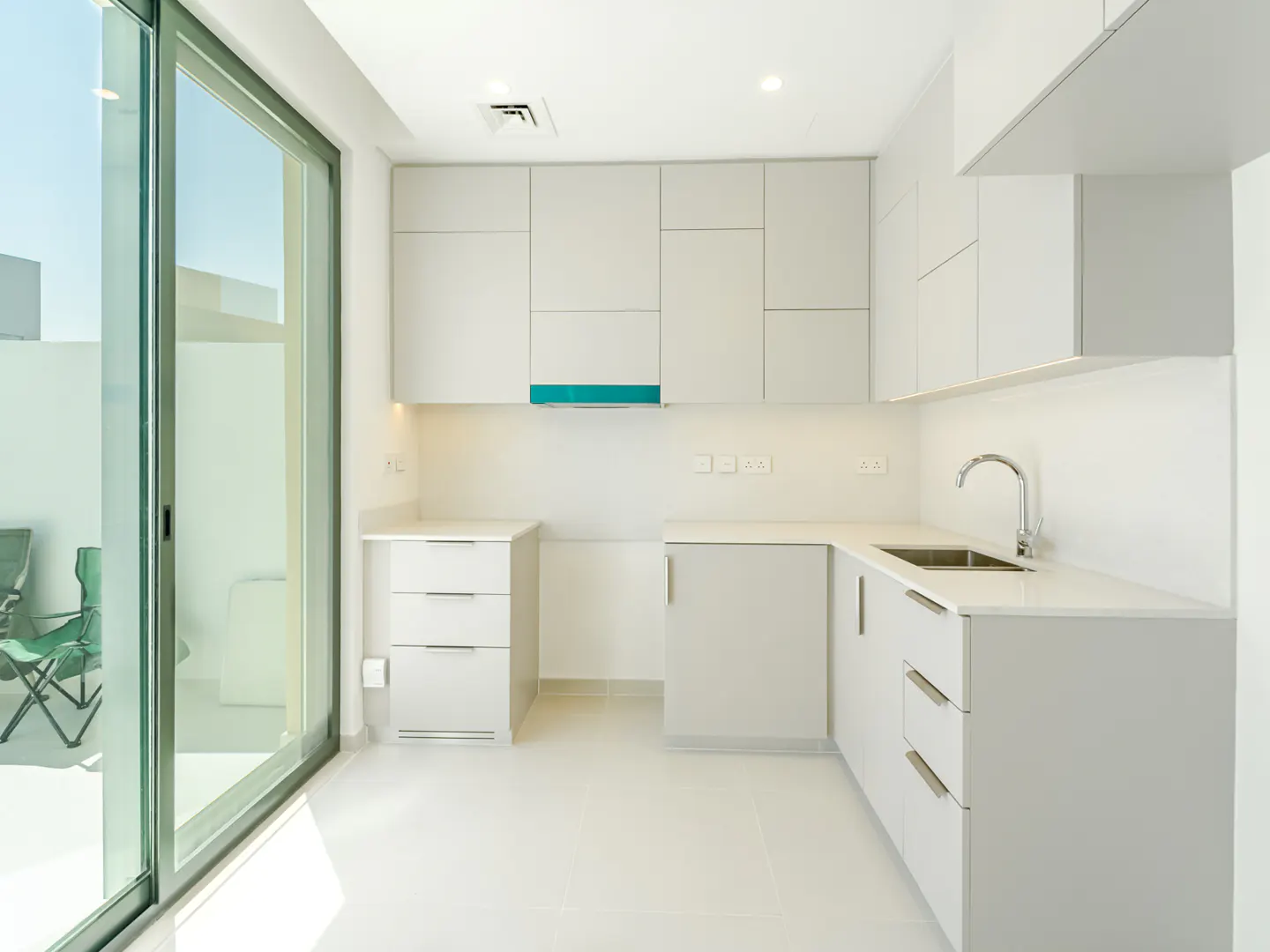 Bright, modern kitchen with white cabinets, countertops, and floor. A large sliding glass door leads to an outdoor patio with green chairs.