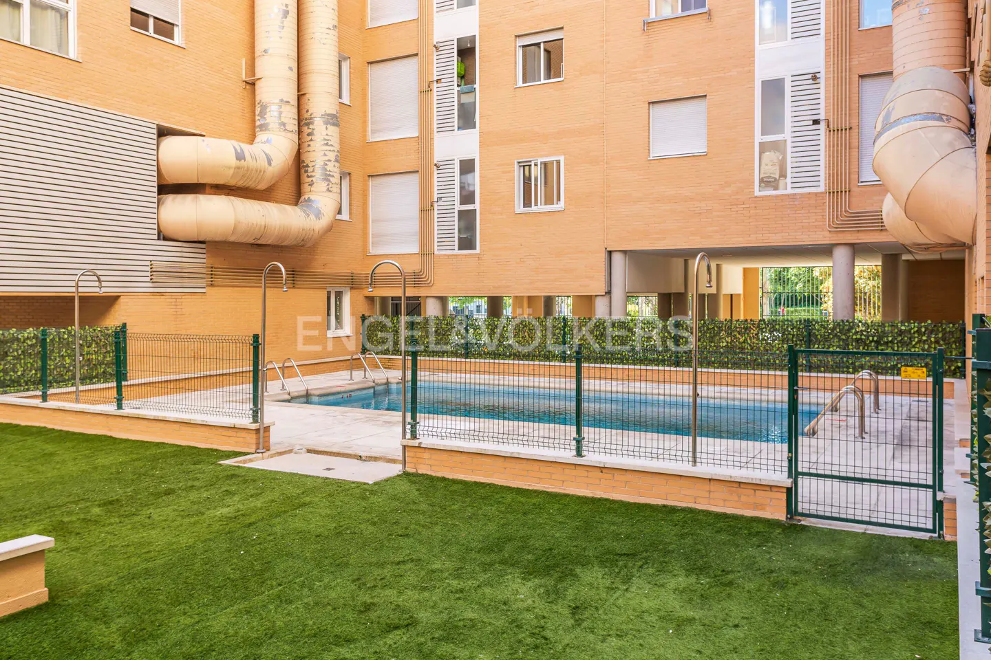 Outdoor pool with green turf and metal fence in a courtyard of a brick apartment building.