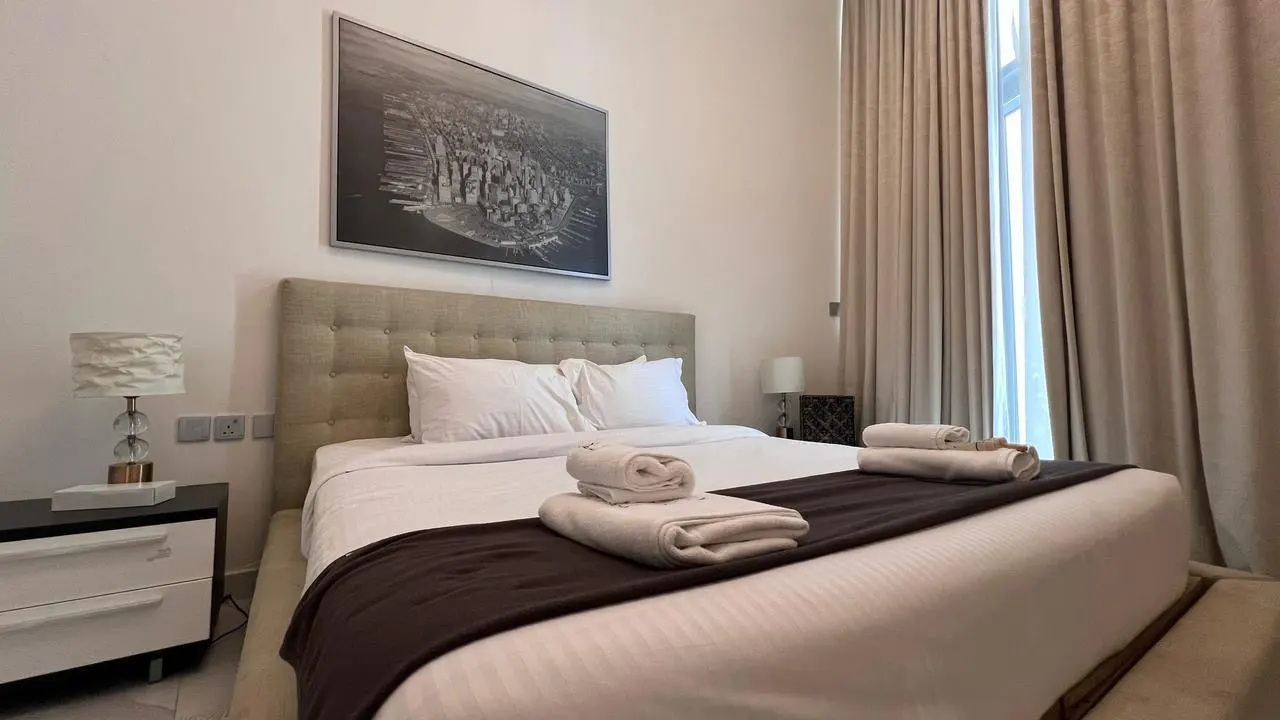 Bedroom with a queen bed, white linens, and a brown throw. A black and white city print hangs above the headboard. Beige curtains frame a window.