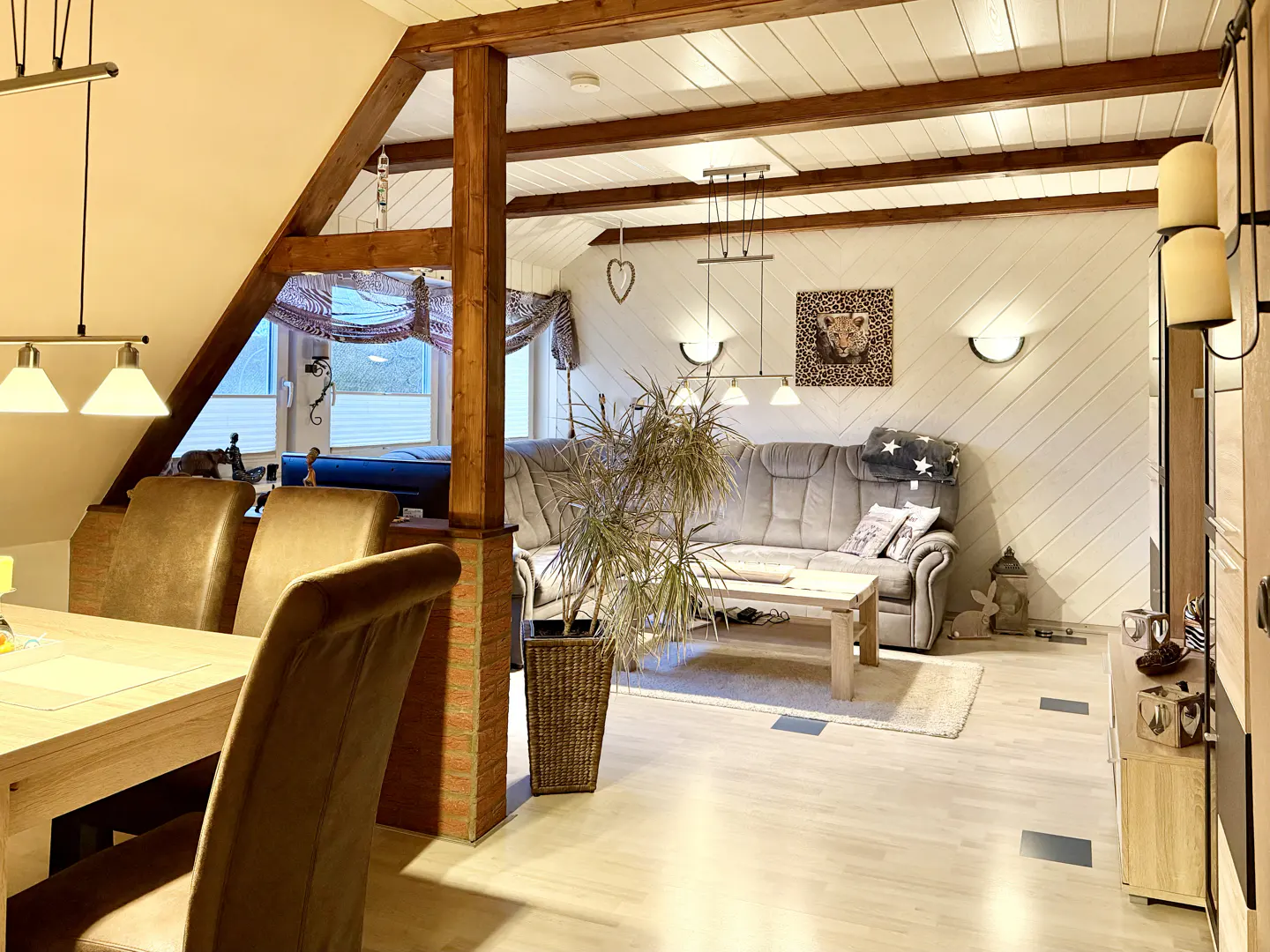 A cozy living room with a gray sofa, wooden beams, and a dining table with brown chairs. A potted plant sits near the sofa.