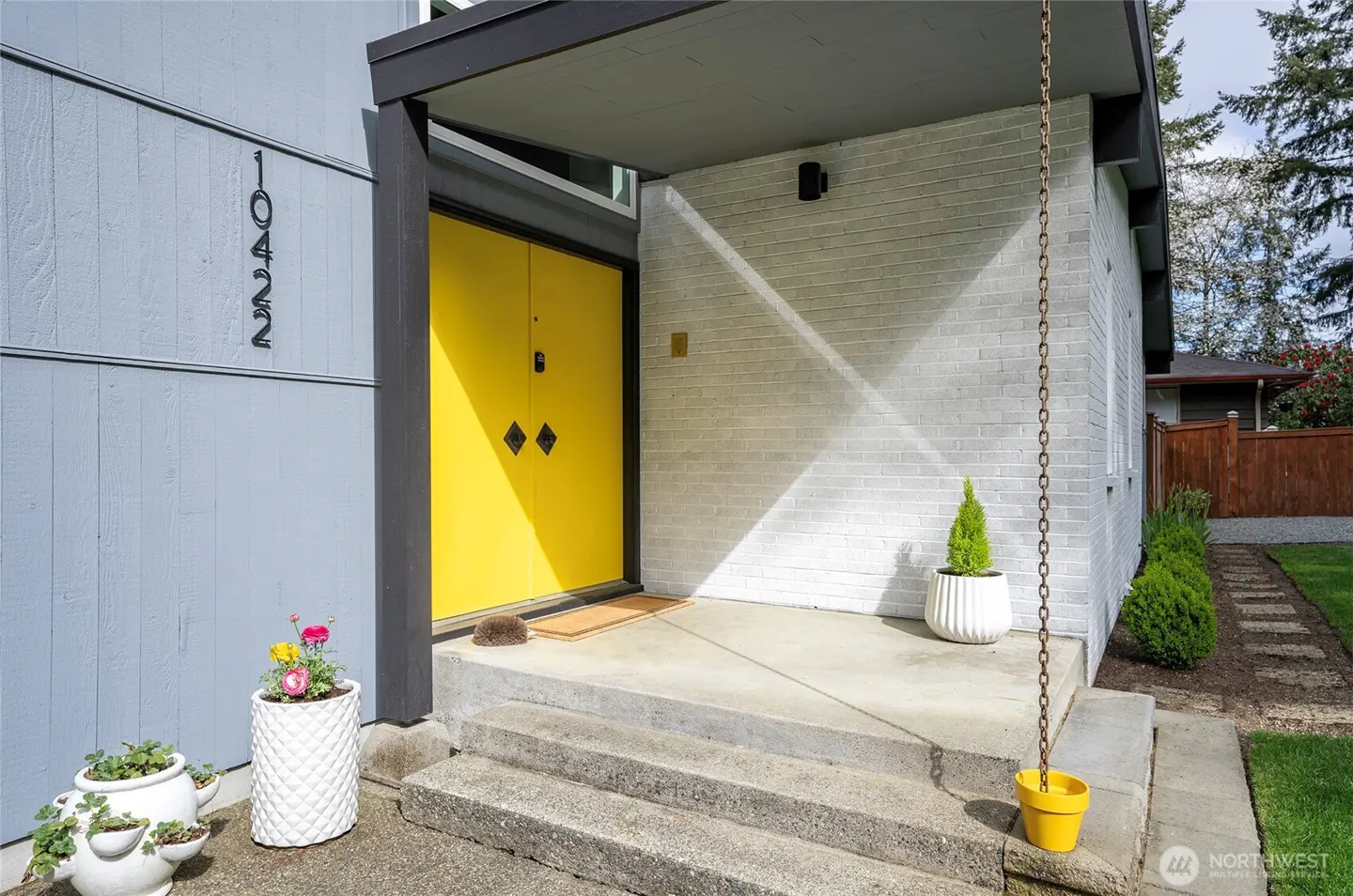 Exterior view of a house with a bright yellow front door, gray siding, and a white brick porch. A rain chain hangs from the porch roof.