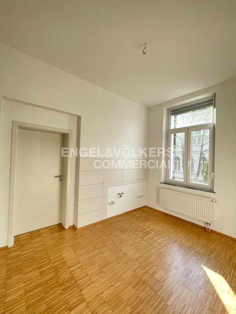 Bright, empty room with wood floors, white walls, a door, and a window with a radiator. Engel & Völkers Commercial logo on the wall.