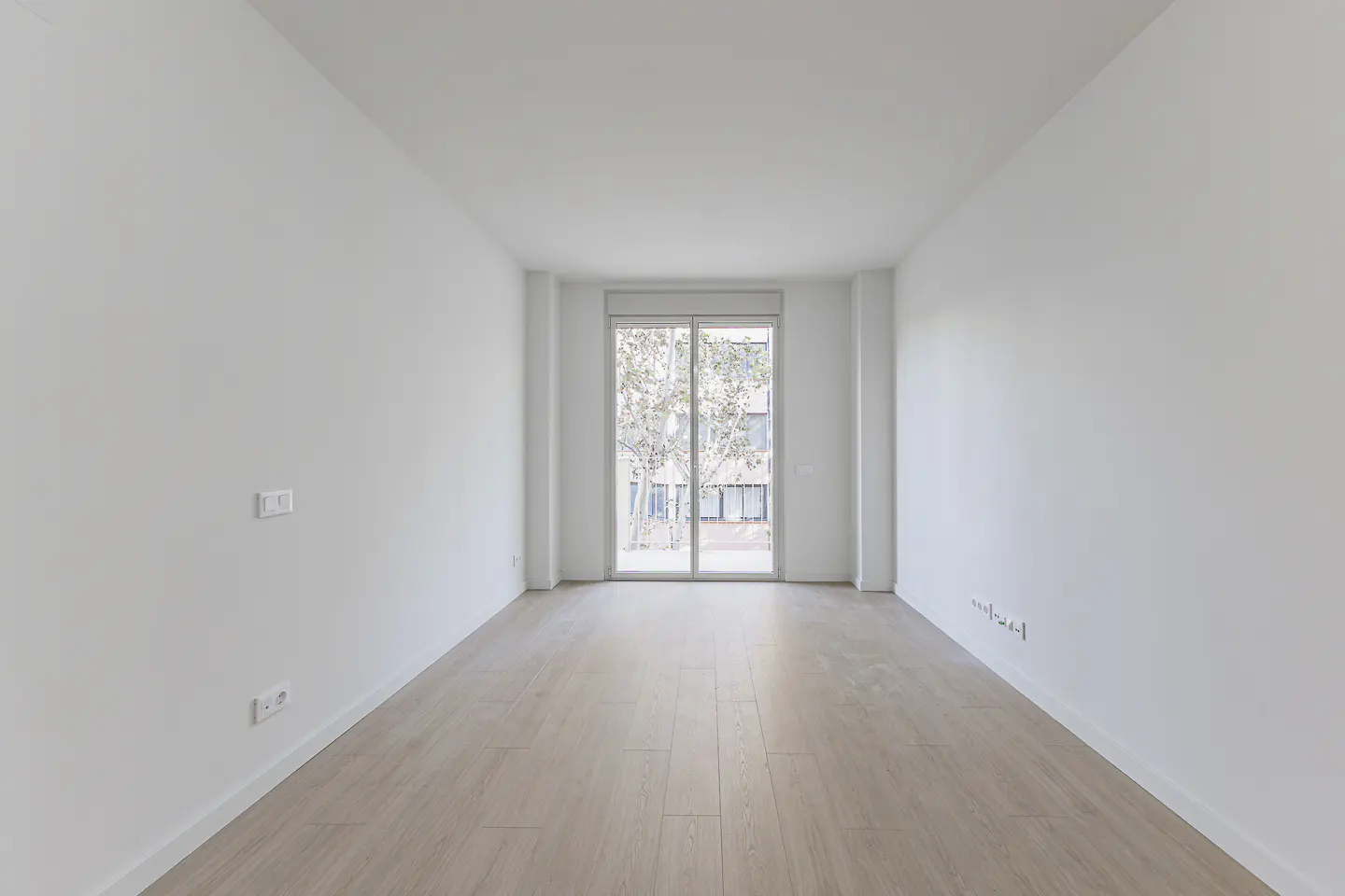 Empty room with light wood floors, white walls, and a sliding glass door to a balcony with a tree outside.