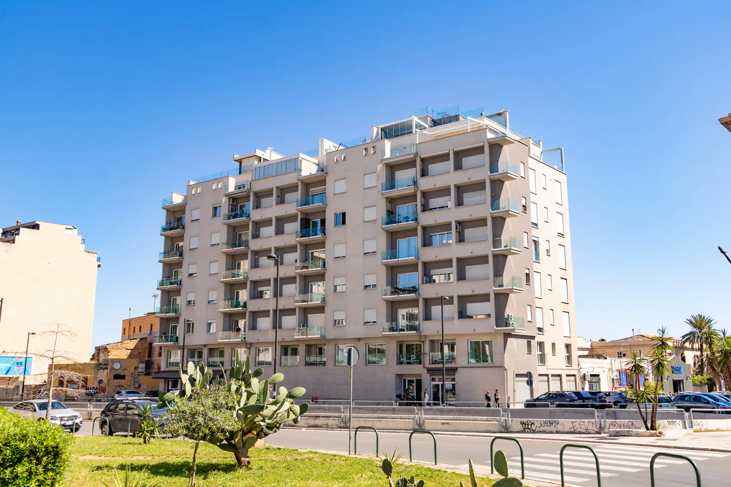 A modern, multi-story apartment building with balconies, set against a clear blue sky. Cars and greenery are visible in the foreground.