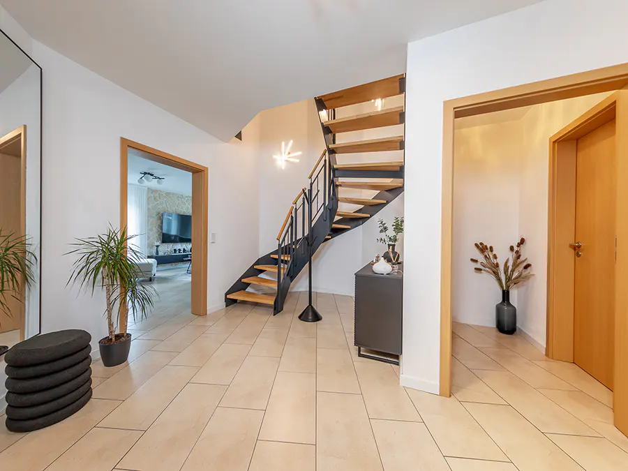 A bright foyer with beige tile floors, a wooden staircase with black railings, and doorways leading to other rooms.