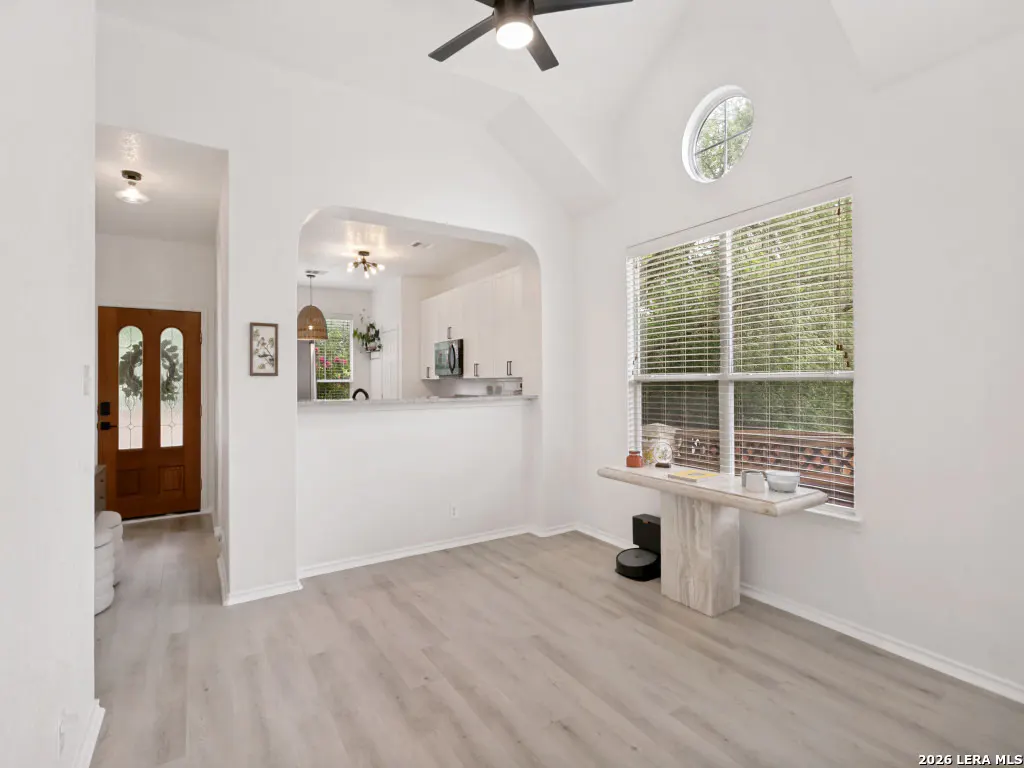 Bright, open-concept living room with white walls, light wood floors, and a vaulted ceiling. A brown front door with a wreath is visible.