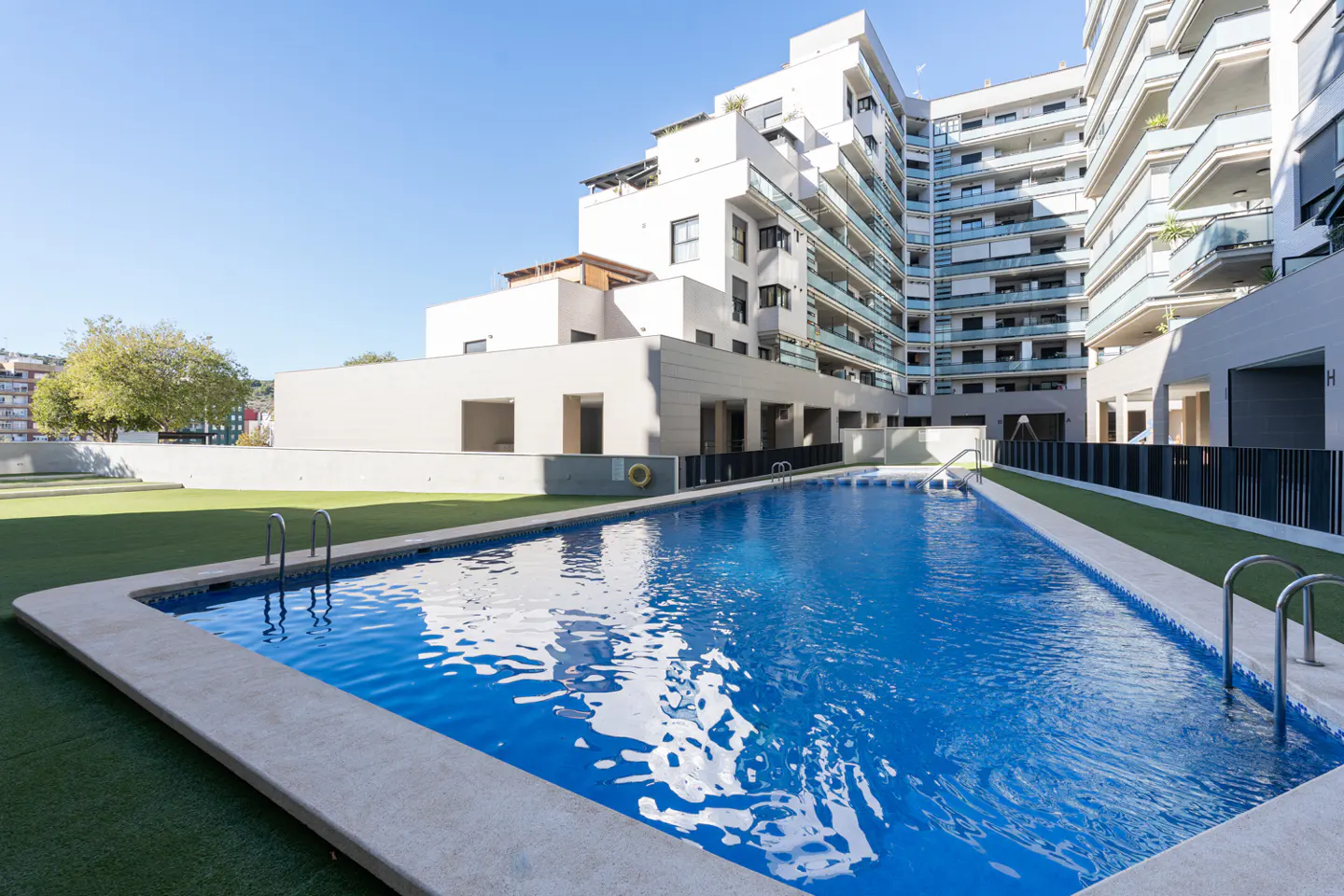 A rectangular swimming pool with blue water sits in front of a modern white apartment building with balconies.