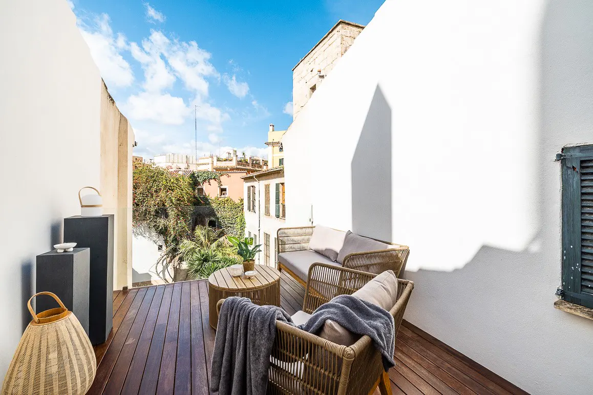 A sunny balcony with wicker furniture, including a chair, loveseat, and table, overlooks a cityscape with lush greenery.