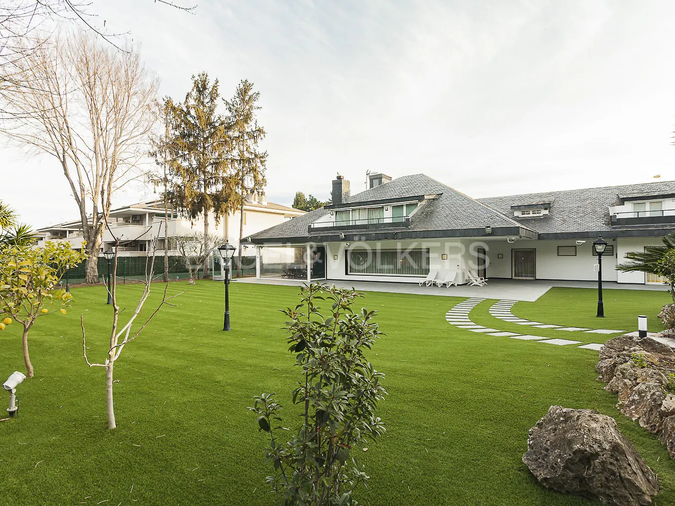 A modern white house with a gray roof, surrounded by a green lawn, trees, and a stone pathway.