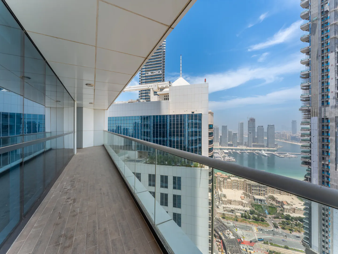 Balcony view of a city skyline with skyscrapers, a bay with boats, and a blue sky with clouds. The balcony has a glass railing and tile flooring.