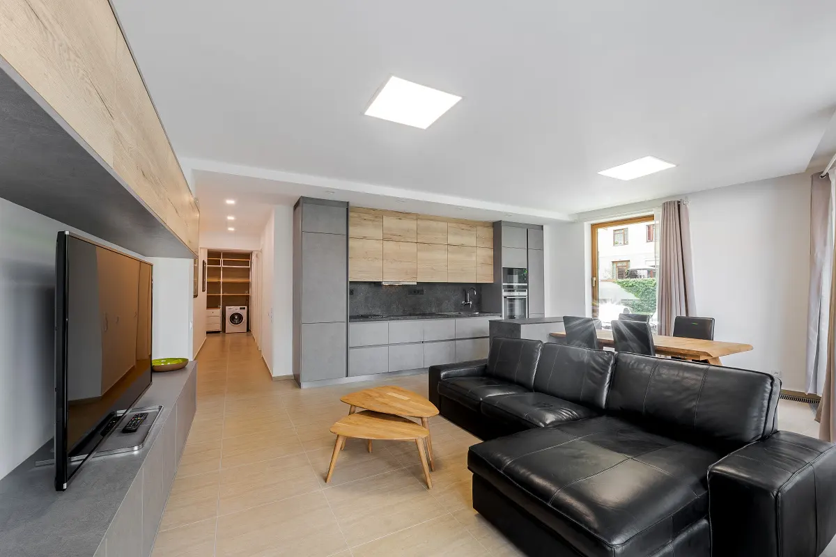 A modern apartment interior with a black leather sofa, wooden coffee tables, and a gray kitchen.
