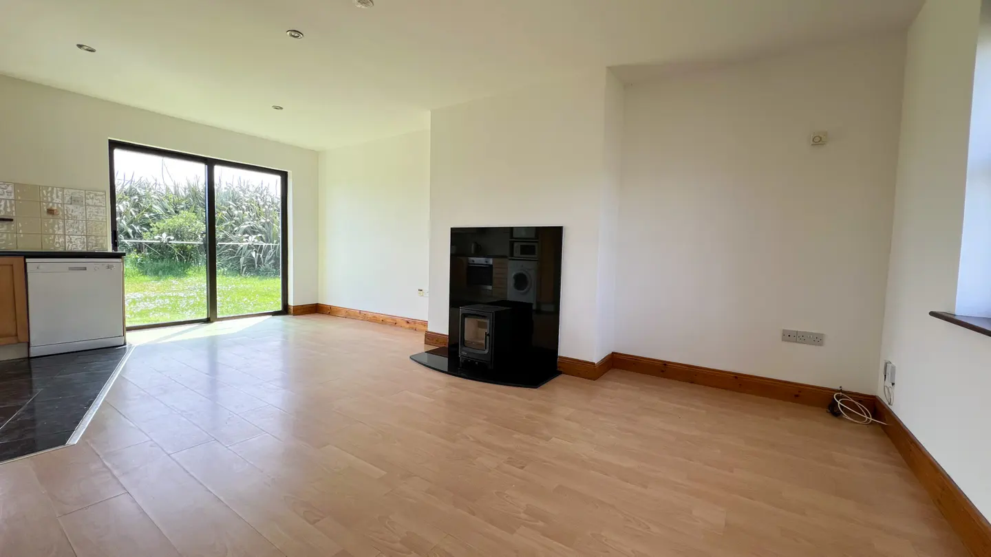Bright, empty room with wood floors, white walls, and a black fireplace. A sliding glass door leads to a green yard. Kitchen appliances are visible.