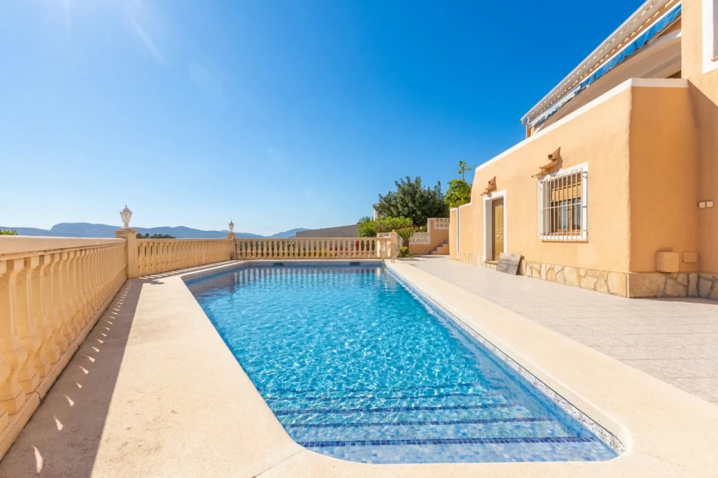 Outdoor pool with clear blue water, tan stone patio, and a peach-colored house under a bright blue sky.