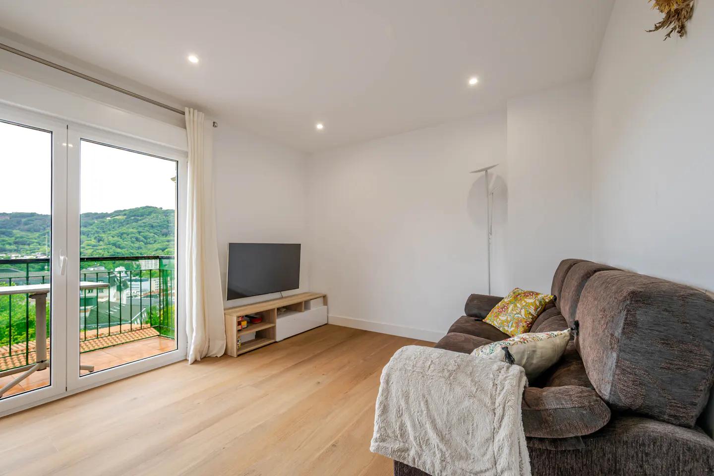Bright living room with wood floors, brown sofa, TV, and balcony doors with a view of green hills.