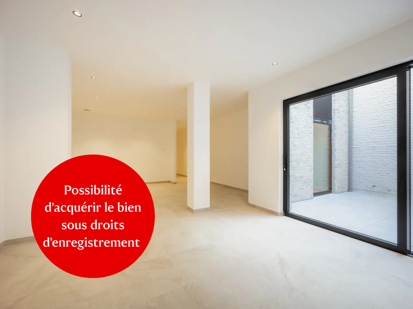 Bright, empty room with white walls, gray floor, and black-framed sliding glass doors to a courtyard. Red circle with white text.