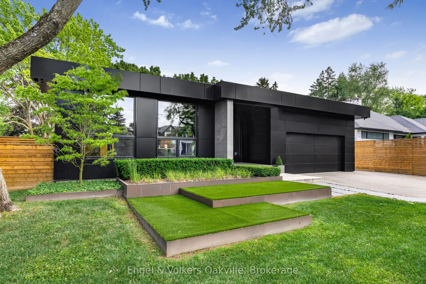 Modern black house with green lawn and tiered turf steps leading to the entrance. Large windows reflect the surrounding trees.