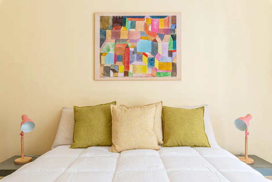 Bedroom with a white bed, green and yellow pillows, pink lamps, and a colorful abstract painting on a yellow wall.