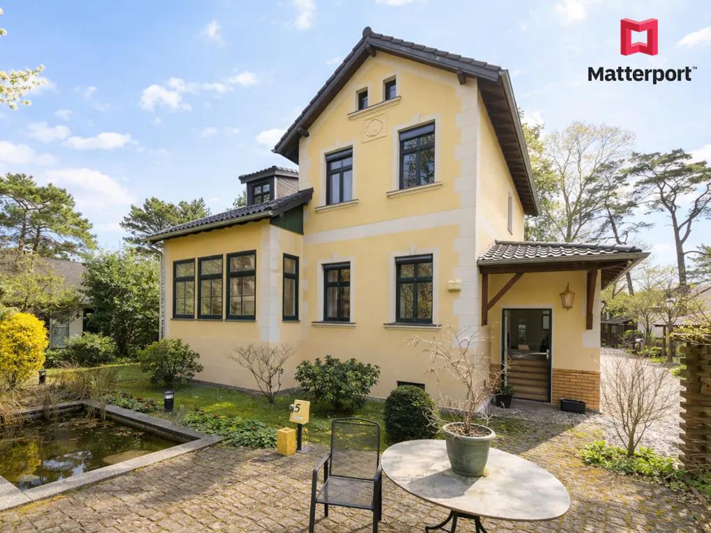 Two-story yellow house with dark trim, a stone patio, a small pond, and outdoor furniture on a sunny day.