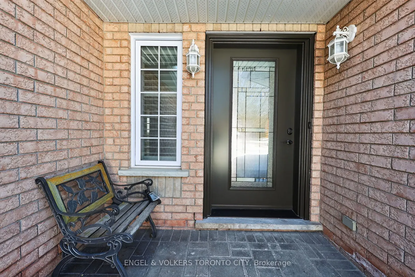 Brick house entrance with a black door, white window, and a bench. Two white sconces flank the door.