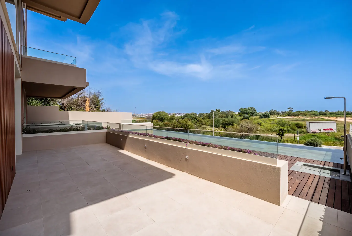 A tan patio with glass railings overlooks green trees under a blue sky.