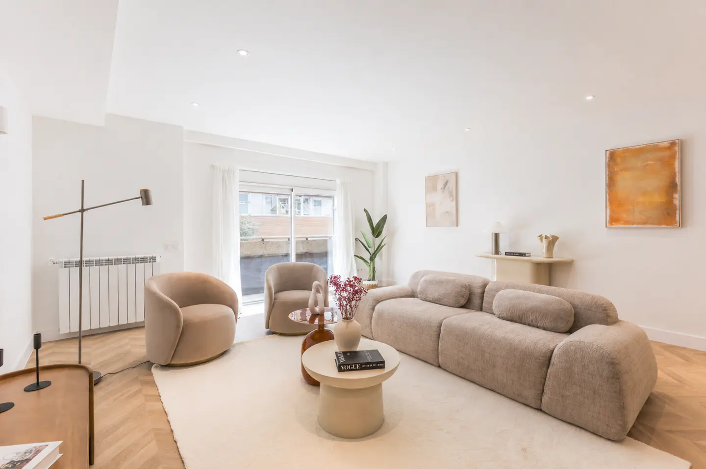 Bright living room with beige sofa, chairs, and white rug. A round coffee table holds books and flowers. Art hangs on the white walls.