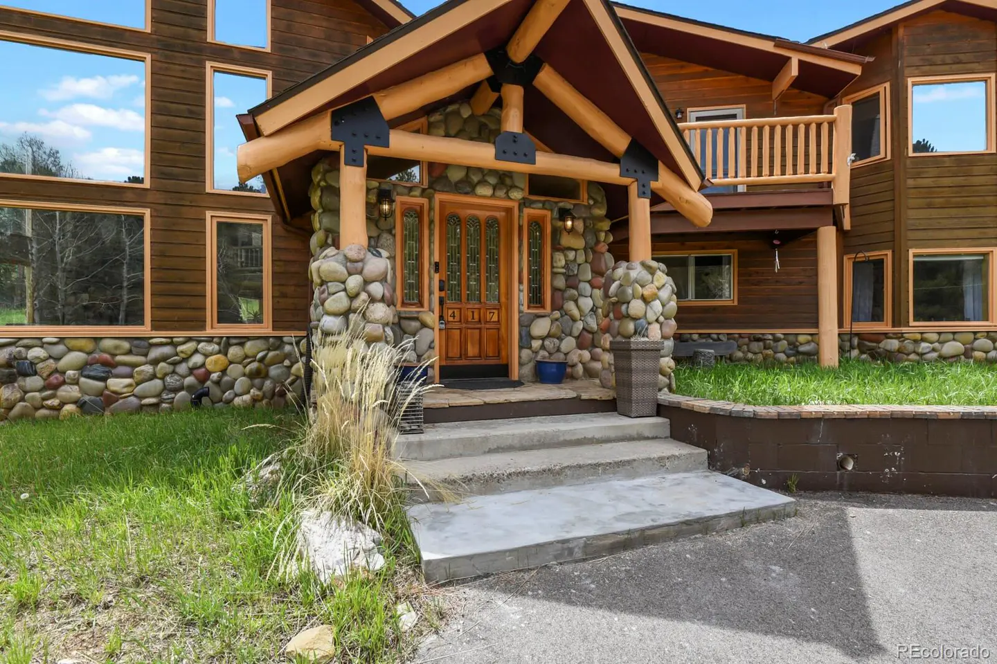 Log cabin home exterior with stone accents. The front door is wood with glass panels and the house number 417. Concrete steps lead to the entrance.