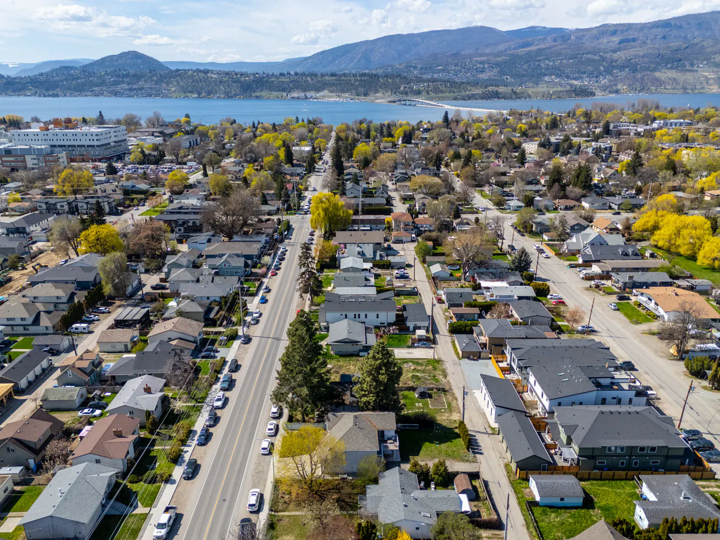 Aerial view of a residential neighborhood with houses, trees, and a road with parked cars. A lake and mountains are visible in the background.