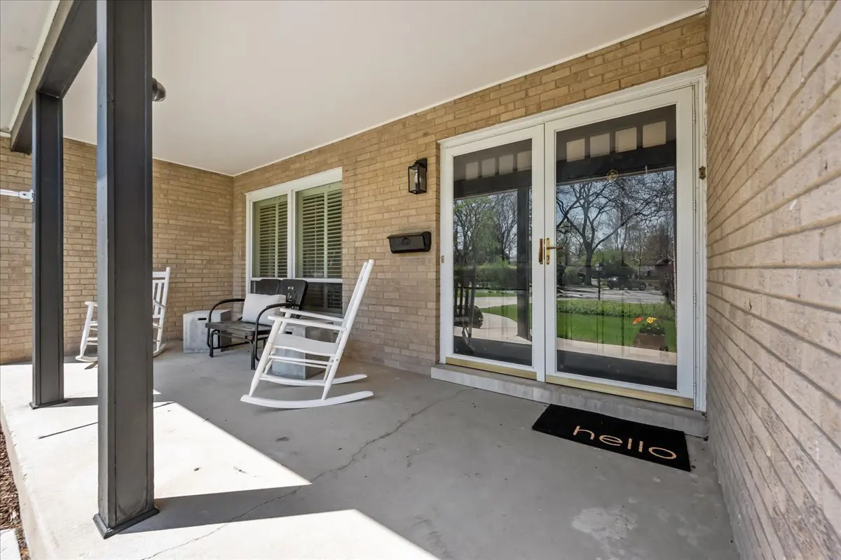 Covered porch with brick walls, white trim double doors, and a "hello" doormat. Rocking chairs and a bench provide seating.