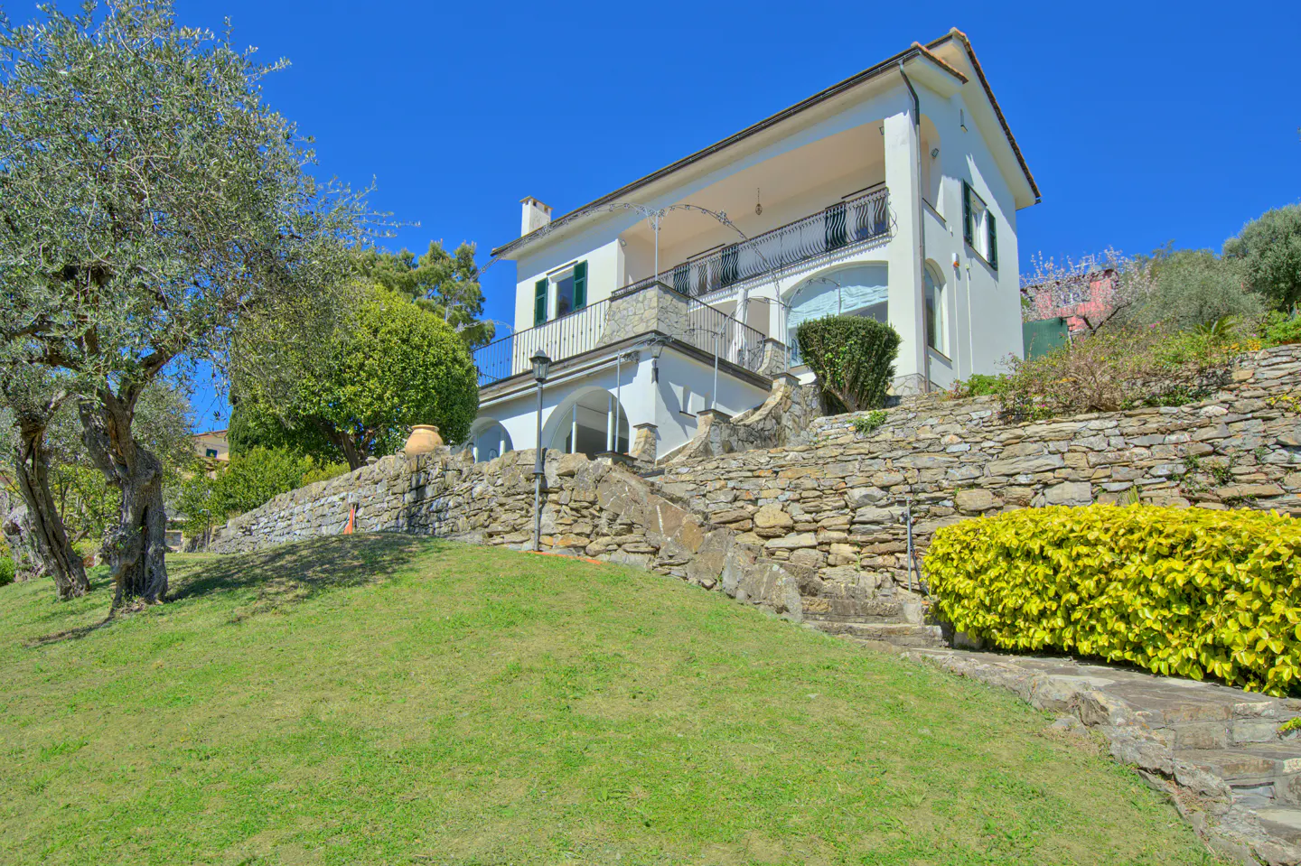 A two-story white house with a balcony sits on a hill with stone walls and green grass. Blue sky above.