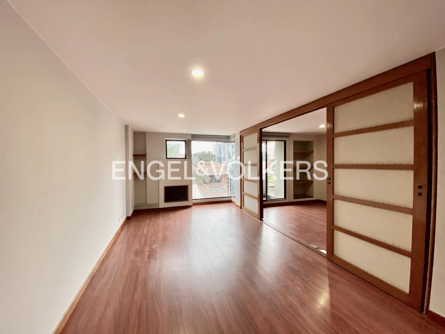 A bright, empty room with wood floors, white walls, and sliding wood doors. A fireplace and window are on the far wall.