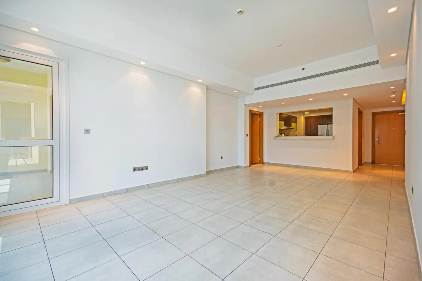 Bright, empty apartment with white walls, tile floor, and a pass-through to the kitchen. A glass door leads to a balcony.
