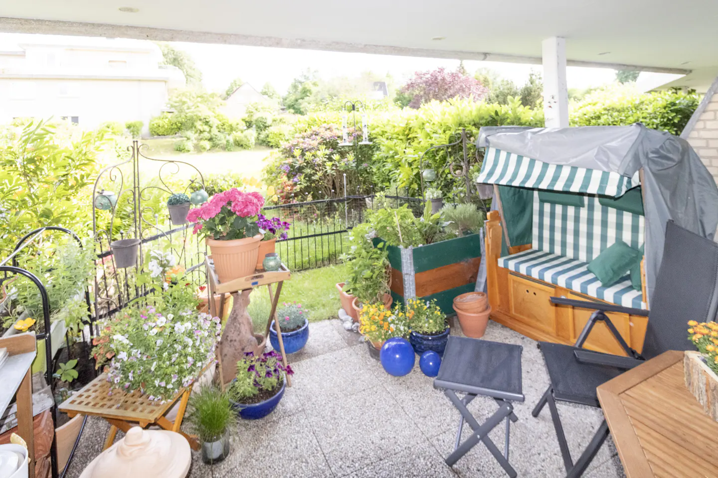 A cozy patio filled with potted flowers, a wooden bench with a green striped canopy, and outdoor seating overlooking a garden.