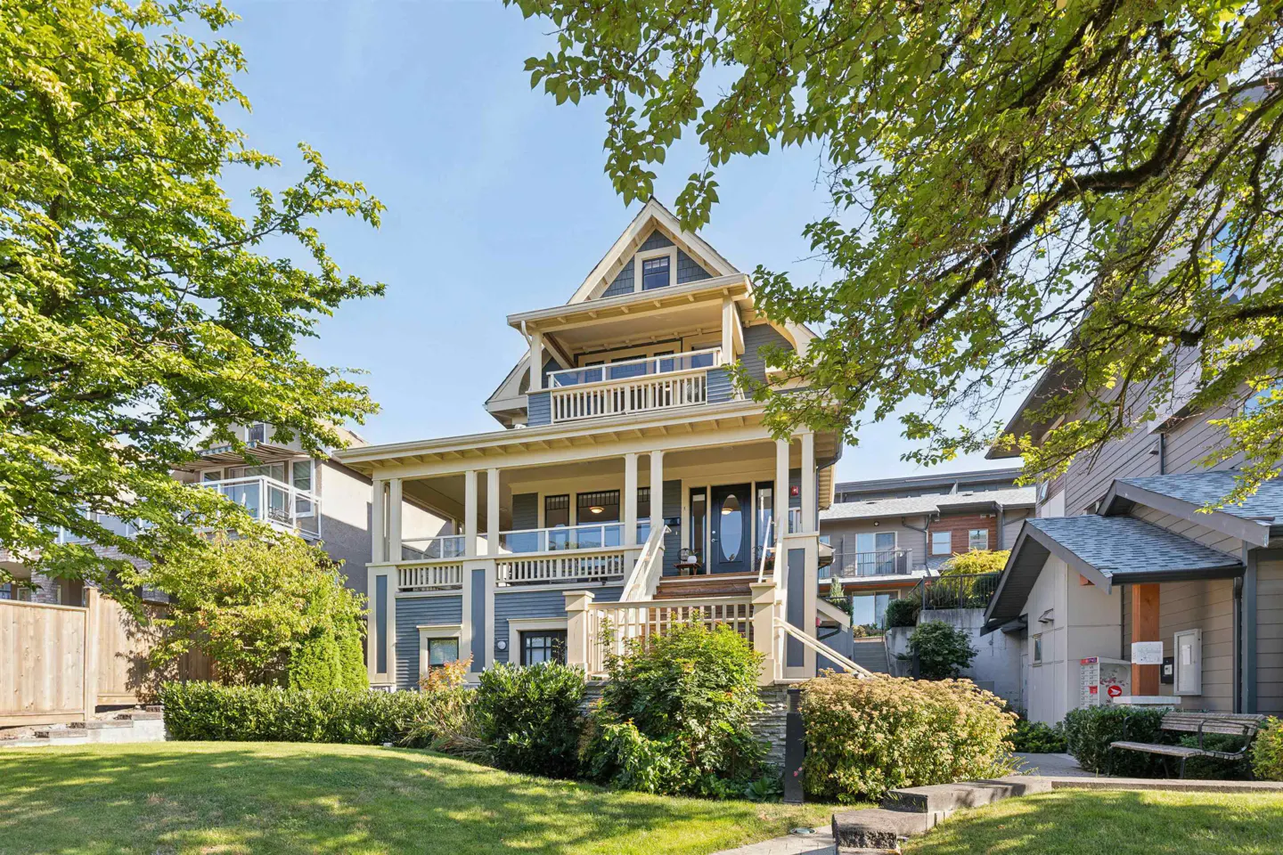 Two-story blue house with white trim, a large porch, and a green lawn. Trees frame the house against a blue sky.