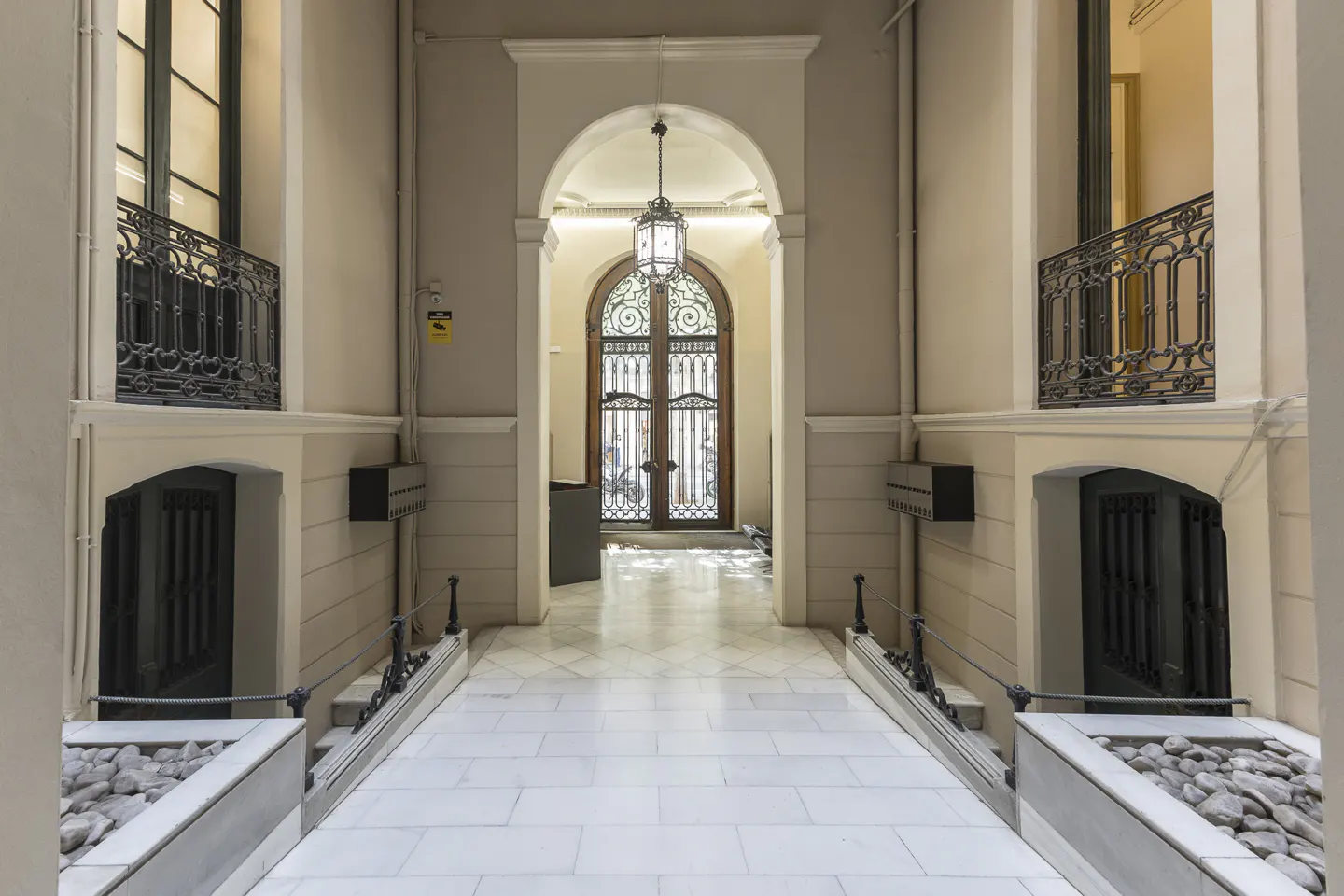 Hallway with white marble floors, arched doorway, and decorative ironwork. A lantern hangs from the ceiling. Balconies with black railings are visible.